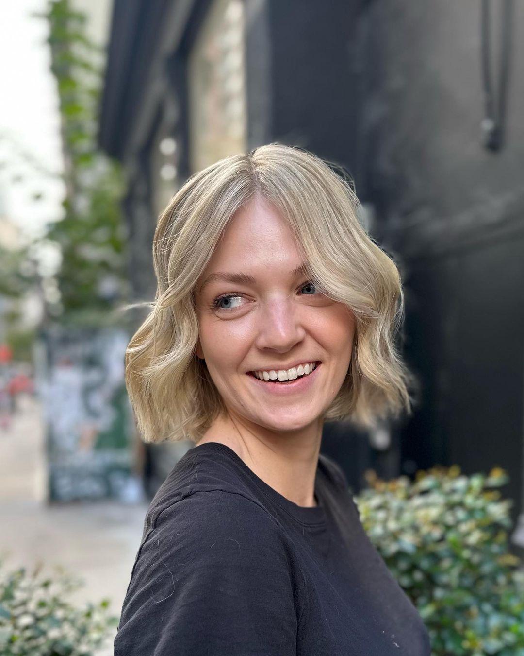 A short, wavy bob cut with platinum blonde and ash blonde hues, featuring a center part and textured ends for a soft, voluminous look.

Color by @simabeautynyc
Cut and style by @juliamannino_hair 3