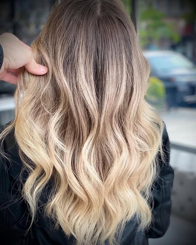Long, wavy hairstyle with a blonde balayage and dark roots, styled in beachy waves.