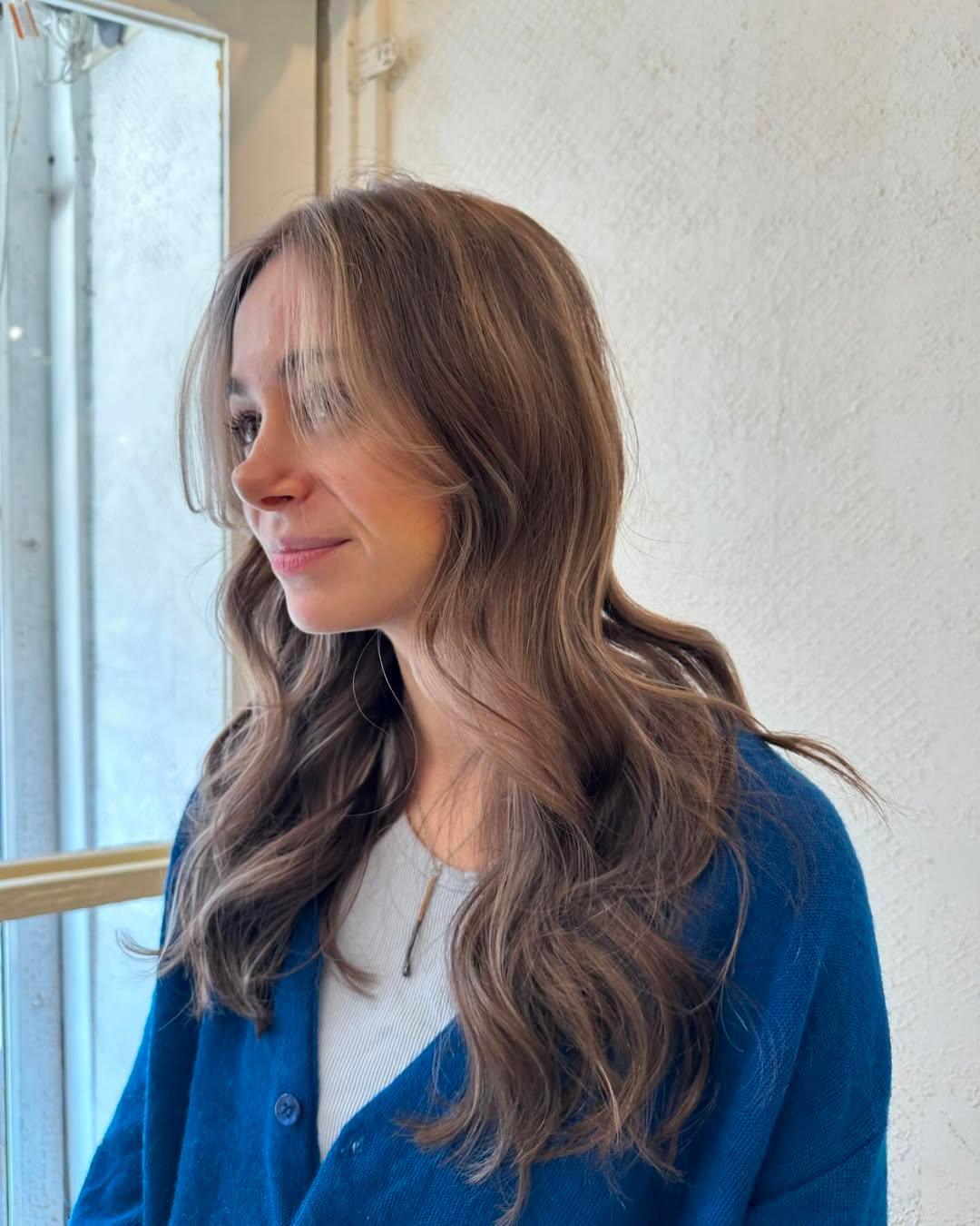 The hairstyle features long, wavy tresses with a blend of light brown and blonde colors, highlighted softly around the face. The layers add dimension and movement to the glossy waves. 2