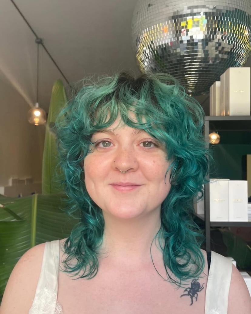 Medium-length, wavy teal hairstyle with shaggy layers and fringe bangs, creating a textured, voluminous look.