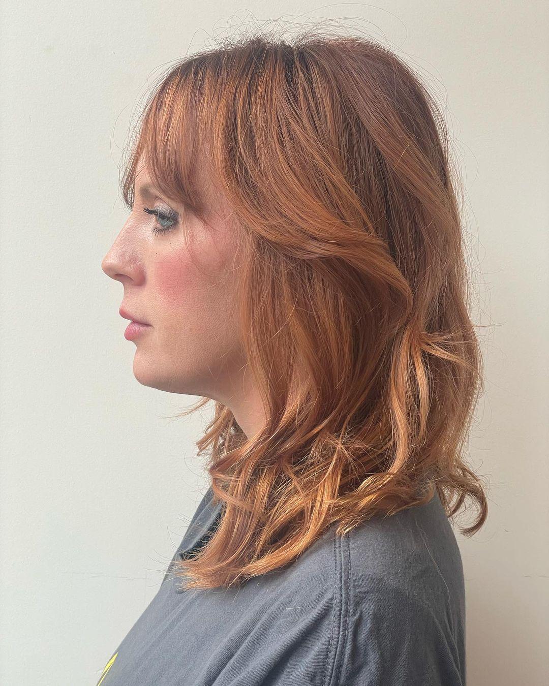 Medium-length, wavy hairstyle in a copper shade, featuring soft waves, long bangs, and a layered, face-framing cut. 2