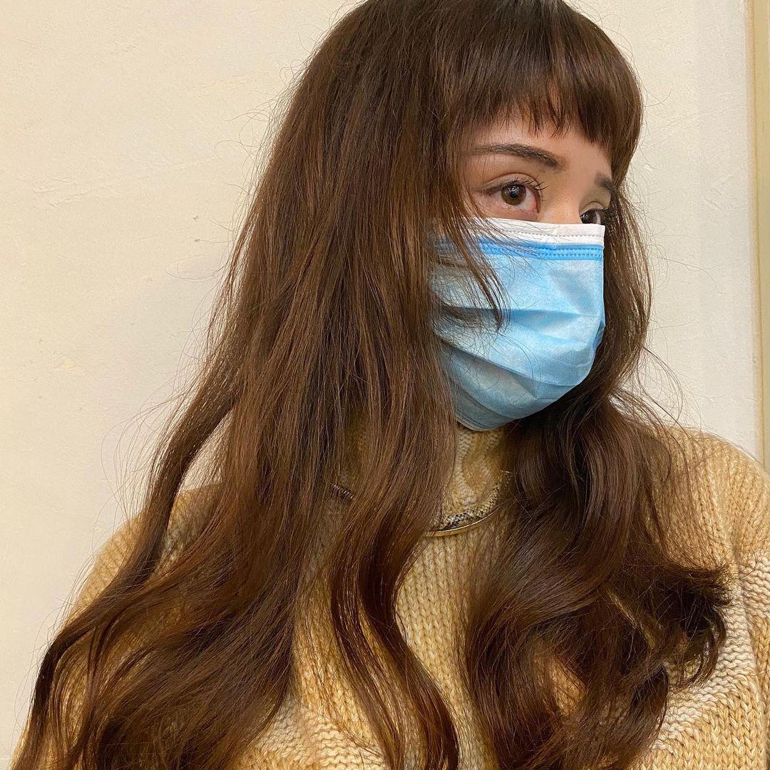 Long chestnut brown wavy hair with micro bangs. The hairstyle features natural, loose waves and layers that add dimension and movement by Coco. 1