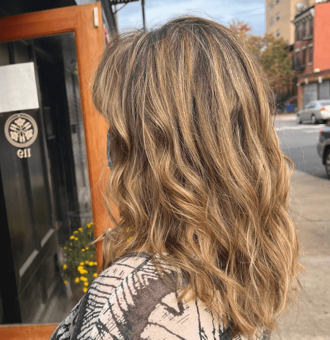 A medium-length wavy hairstyle with face-framing layers and a blended bronde color achieved through balayage. by Salon Mizuno