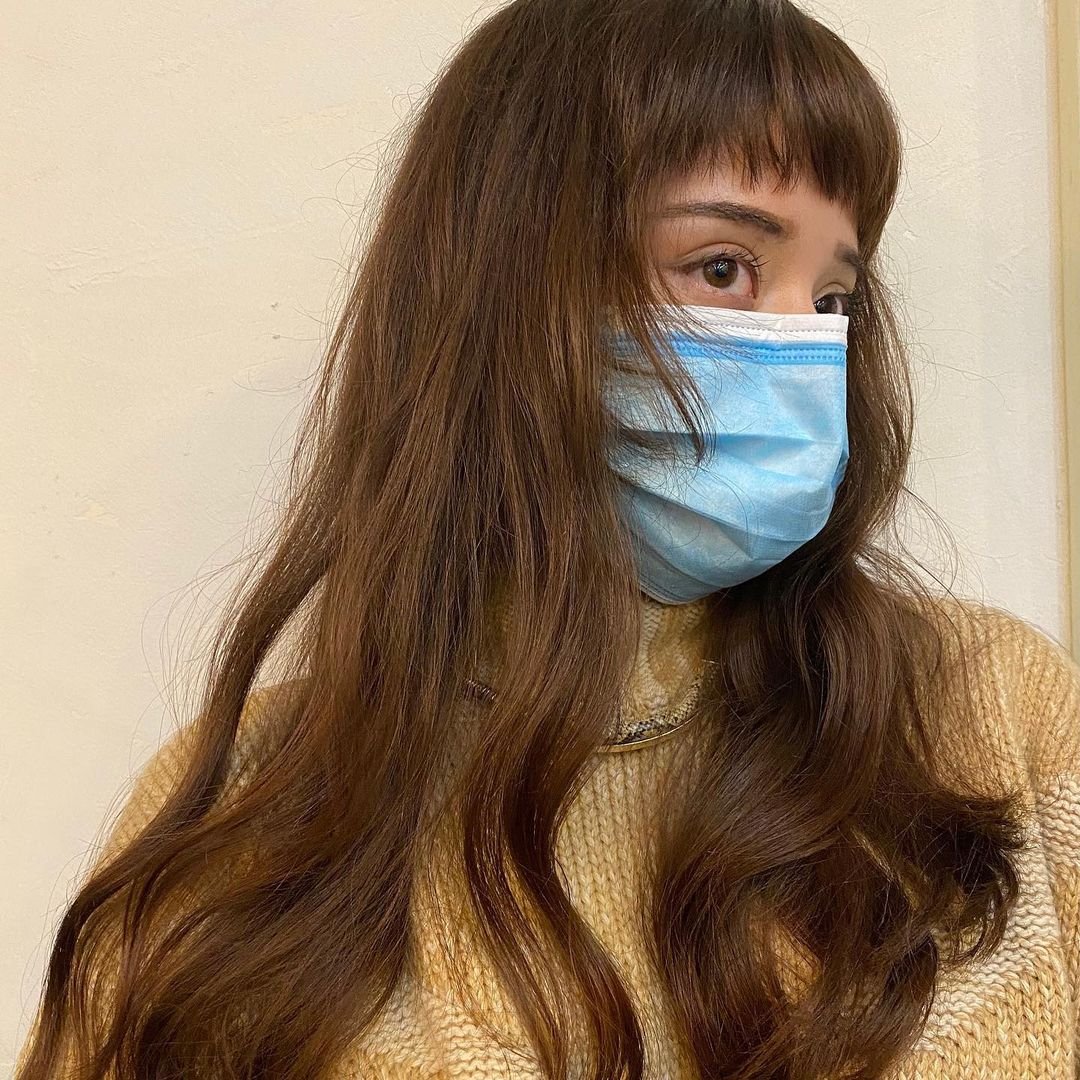 Long chestnut brown wavy hair with micro bangs. The hairstyle features natural, loose waves and layers that add dimension and movement by Coco. 1